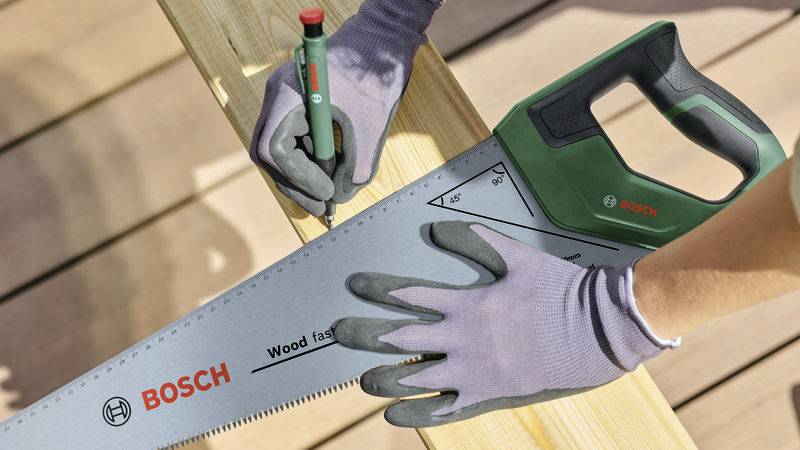 Een persoon met handschoenen meet een houten plank met een Bosch zaag en potlood, blijkbaar voor een doe-het-zelf project.
