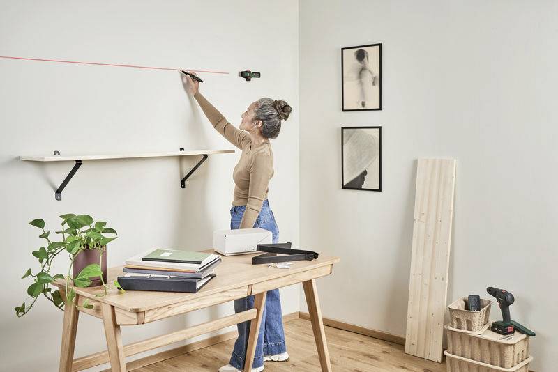 Een vrouw gebruikt een lasergereedschap om een lijn op de muur te markeren. In de ruimte staan een tafel met boeken en een plank.
