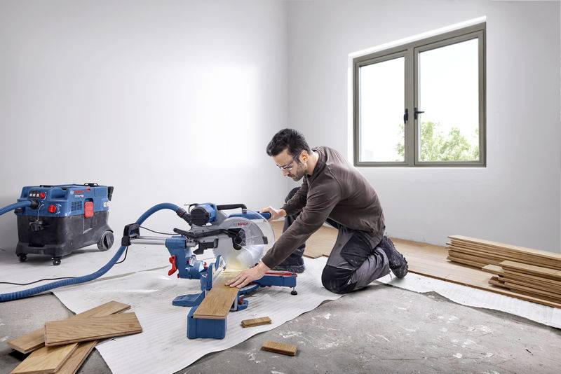 Man zaagt houten plank met een afkortzaag in een ruimte. Stofzuiger aangesloten, houten stukken gestapeld. Raam op de achtergrond.
