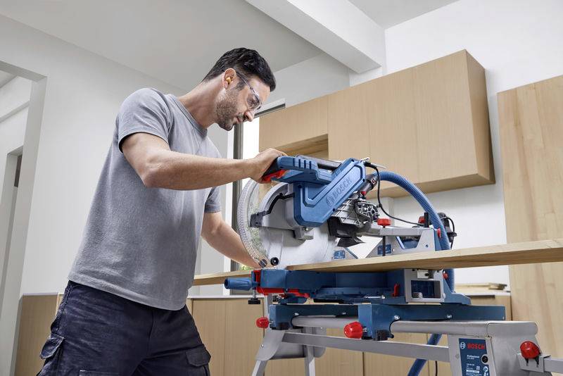 Een man werkt in een werkplaats met een cirkelzaag om een houten plank te zagen. Hij draagt een veiligheidsbril.