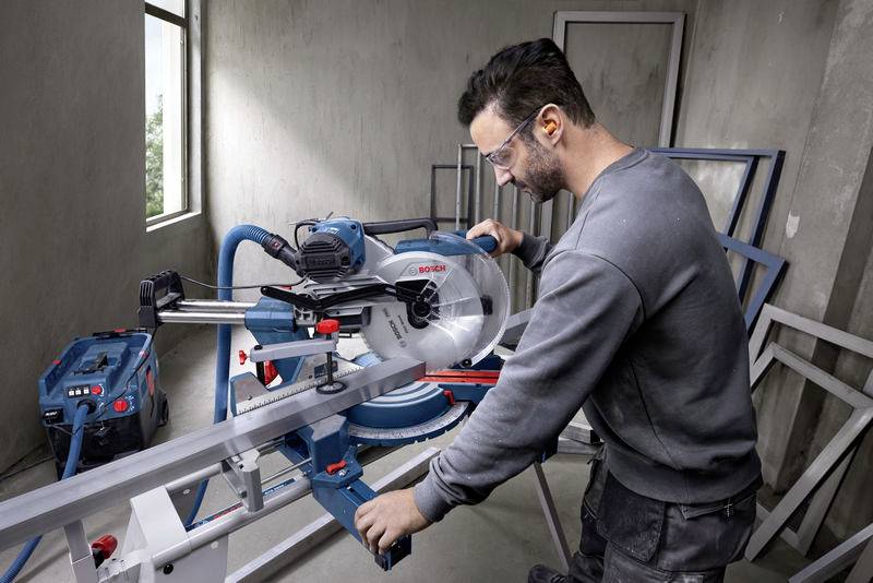 Een man bedient een cirkelzaag in een werkplaats. Hij draagt een veiligheidsbril en gehoorbescherming. De omgeving oogt industrieel en modern.