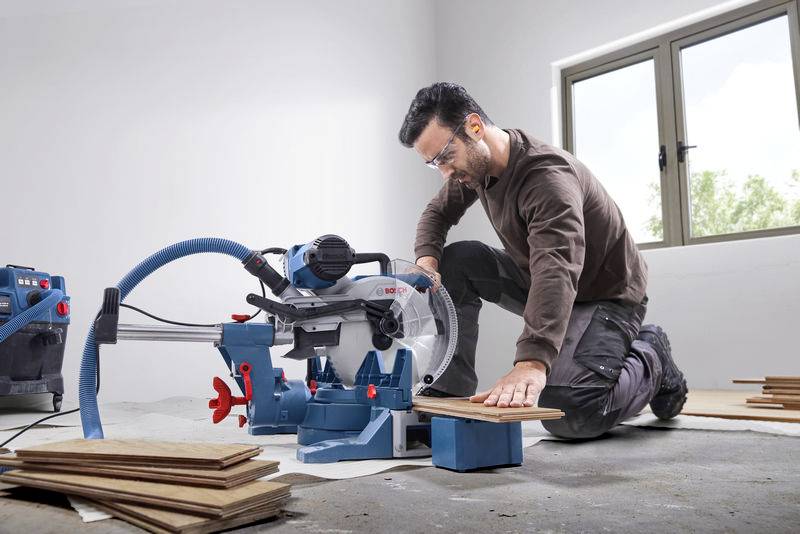 Een man knielt op de grond en zaagt hout met een elektrische tafelzaag in een lichte ruimte. Hij draagt een veiligheidsbril.