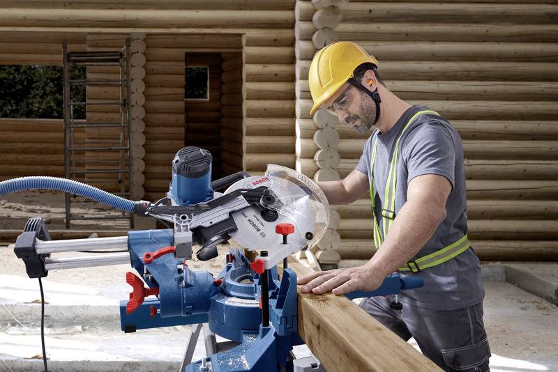 Een bouwvakker met veiligheidshelm en bril zaagt een houten plank op een bouwplaats. Houten gebouwen zijn zichtbaar op de achtergrond.