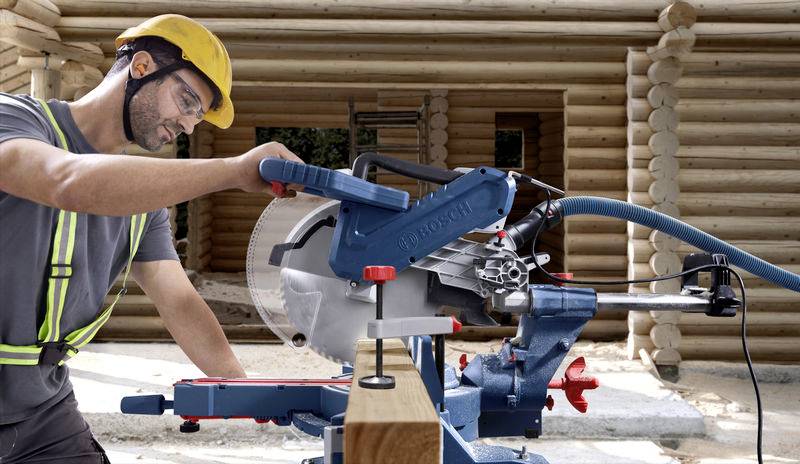 Een bouwvakker met veiligheidshelm en veiligheidsbril gebruikt een tafelcirkelzaag op een houten bouwplaats. Op de achtergrond zijn houten balken zichtbaar.