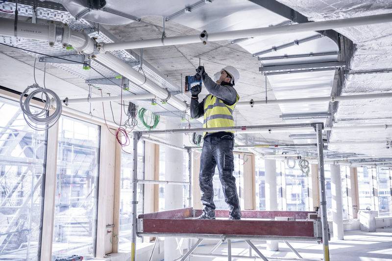 Een bouwvakker staat op een steiger in een gebouw, draagt een veiligheidshesje en helm, en boort in het plafond.