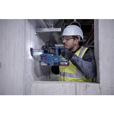 Een bouwvakker draagt een veiligheidshelm en een veiligheidshesje terwijl hij met een elektrische boormachine een gat boort in een betonnen muur.