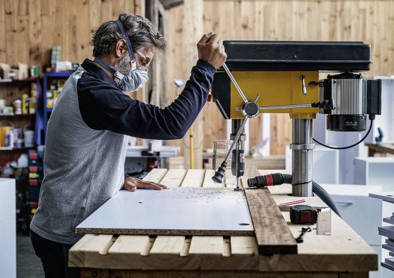 Een man met een masker bewerkt hout met een staande boormachine in een werkplaats.