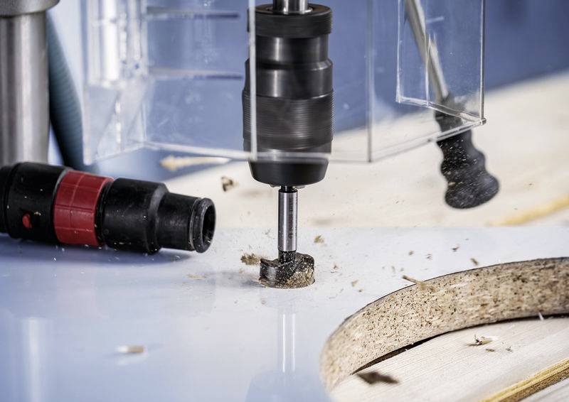 Een boor bewerkt een houten plaat, houtkrullen vliegen. Concentratie op precisiewerk bij het zagen. Gereedschap en materiaal in focus.