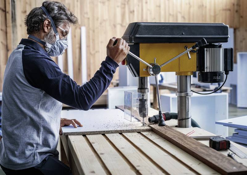 Een man met veiligheidsbril en masker bedient een stationaire houtboormachine in een werkplaats, geconcentreerd op zijn werk.