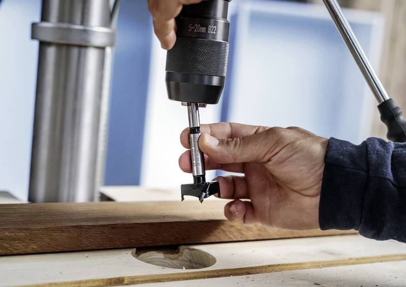 Een hand houdt een houten boor vast onder een boormachine, boven een stuk hout, klaar om een gat te boren.