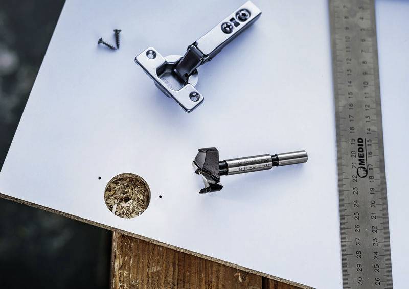 Een metalen scharnier, een boor en een schroef liggen naast een plank met een geboord gat. Rechts ligt een metalen hoekprofiel.