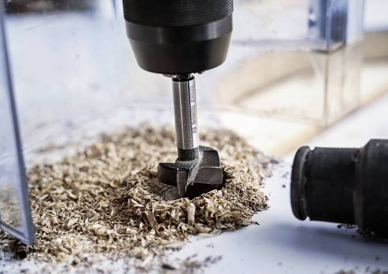 Een boor maakt een rond gat in een houten plank, omgeven door houtspaanders. De boorkopper is zilverkleurig en cilindrisch, met een zwarte handgreep.