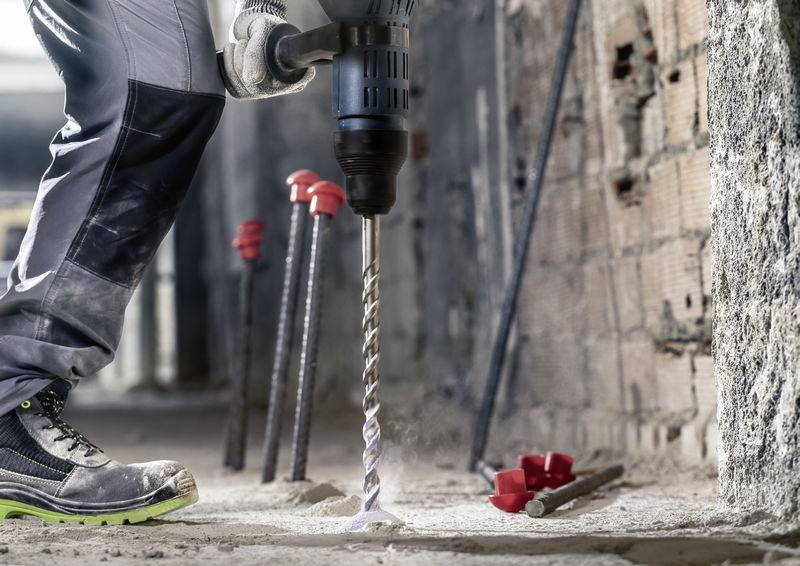 Een persoon boort met een elektrische boormachine in een betonnen muur. Op de achtergrond zijn stalen staven en rode doppen zichtbaar.
