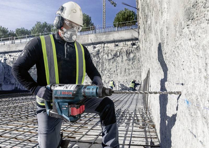 Een bouwvakker met veiligheidshelm en mondmasker boort met een elektrische boormachine in een betonnen muur op een bouwterrein.