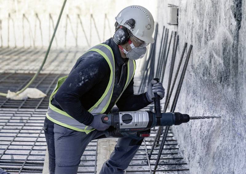 'Bouwvakker boort in betonwand. Draagt veiligheidshelm, vest en masker. Bouwplaats met stalen versterkingen op de achtergrond.'