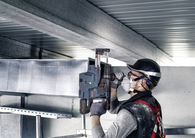 Een bouwvakker met helm en veiligheidsbril bevestigt een metalen constructie aan het plafond met een boormachine in een industriële omgeving.