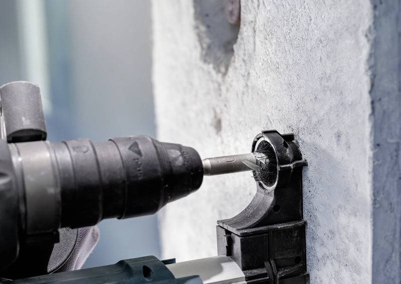 Een persoon boort een gat in een betonnen muur met een elektrische boormachine. De boormachine is dicht bij een kunststof boorbit gepositioneerd.
