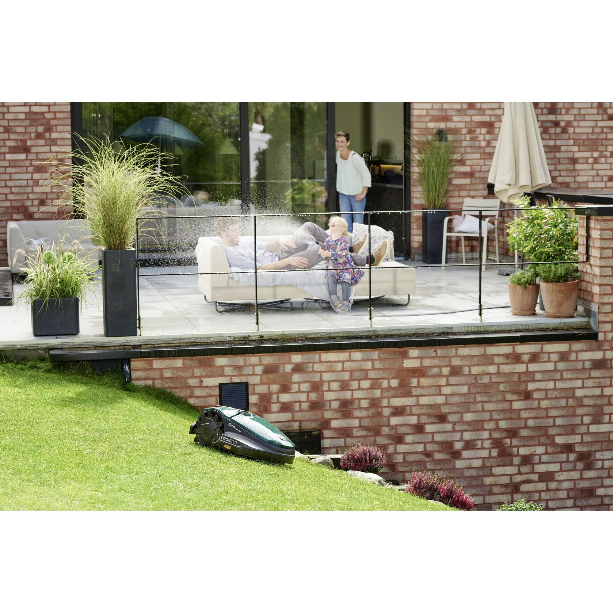 Een gezin geniet van het terras; een robotmaaier rijdt over het gazon. Op de achtergrond staan planten en een parasol.