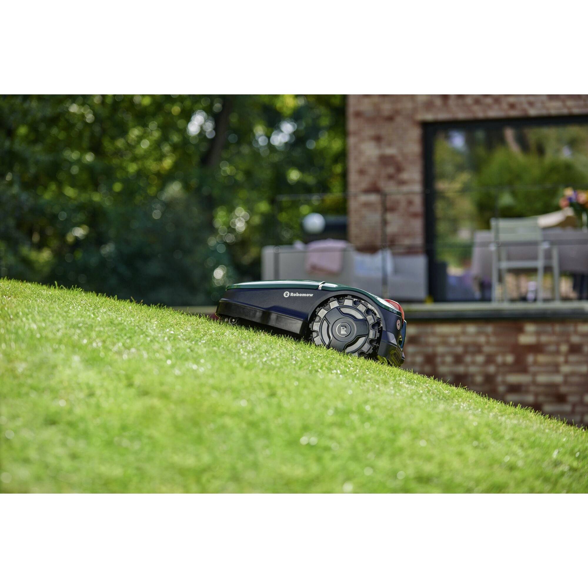 Een robotmaaier rijdt een met gras begroeid talud op, omringd door bomen en een bakstenen gebouw op de achtergrond.