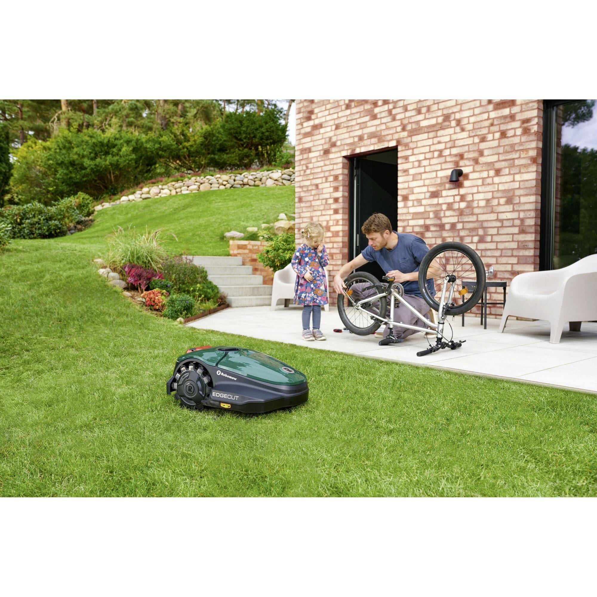 Een kind en een volwassene repareren een fiets op een terras. In de tuin maait een robotmaaier het gras.