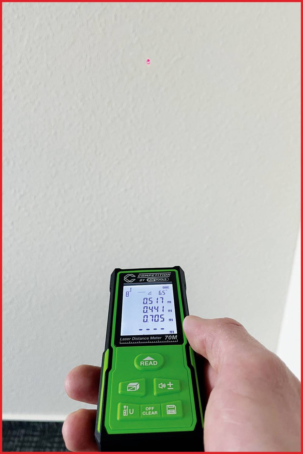 Een hand houdt een groene laser-afstandsmeter vast. Een rode laserpunt is zichtbaar op een witte muur. Het apparaat toont de afstandsmeting.