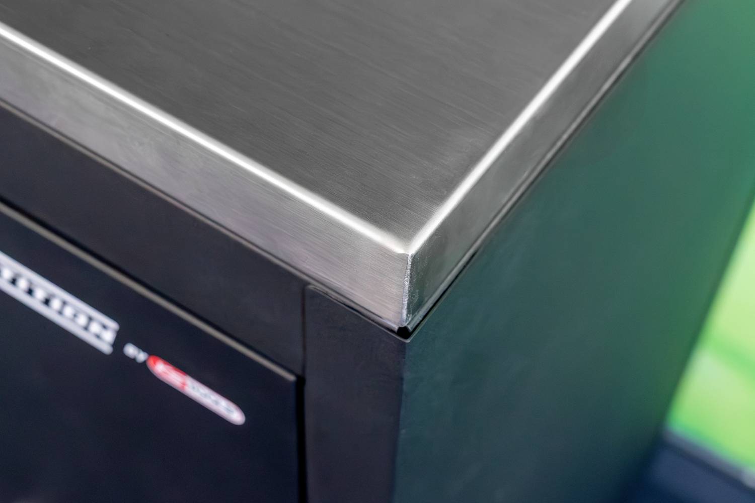 Close-up van een zwarte metalen doos met een glanzende roestvrij stalen rand. De hoek toont het gladde oppervlak en het robuuste materiaal.