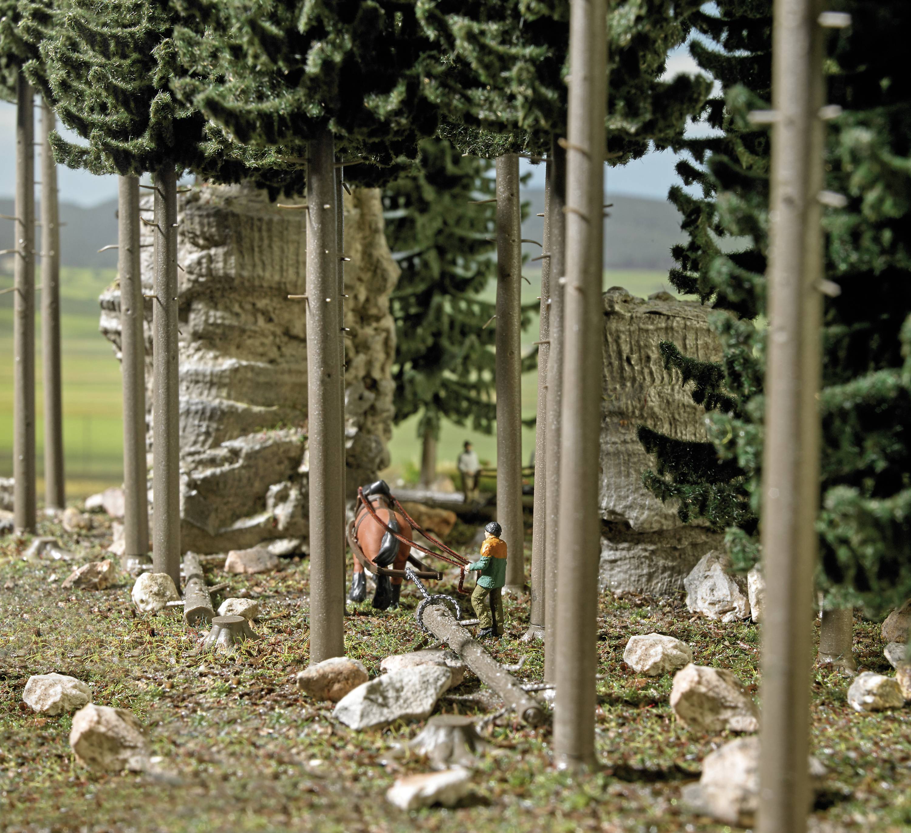 'Miniatuurscène van een bos met twee figuren die boomstammen doorzagen. Hoge bomen en rotsen op de achtergrond.'