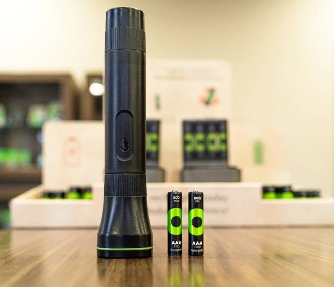 Een zwarte zaklamp staat rechtop op een tafel, omringd door twee AAA-batterijen. Op de achtergrond zijn wazig schappen zichtbaar.