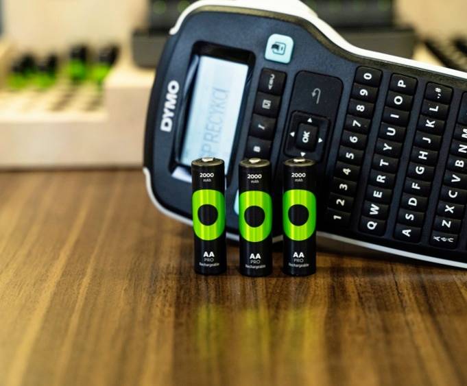 Een etikettenprinter en drie oplaadbare AA-batterijen op een houten tafel. De printer heeft een display en een toetsenbord.