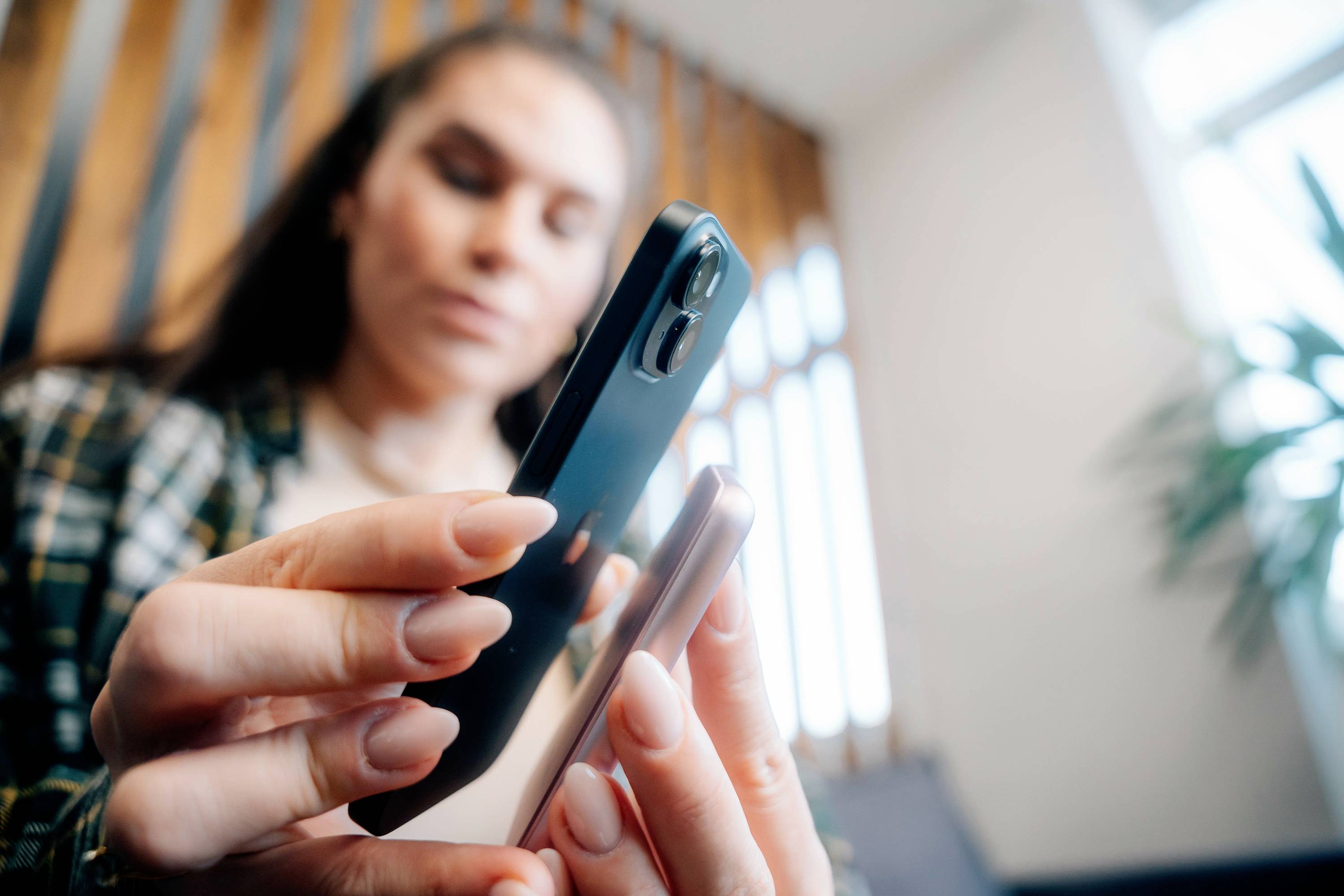 Een vrouw houdt en bekijkt twee smartphones in haar handen. Op de achtergrond zijn houten balken en onscherpe planten te zien.