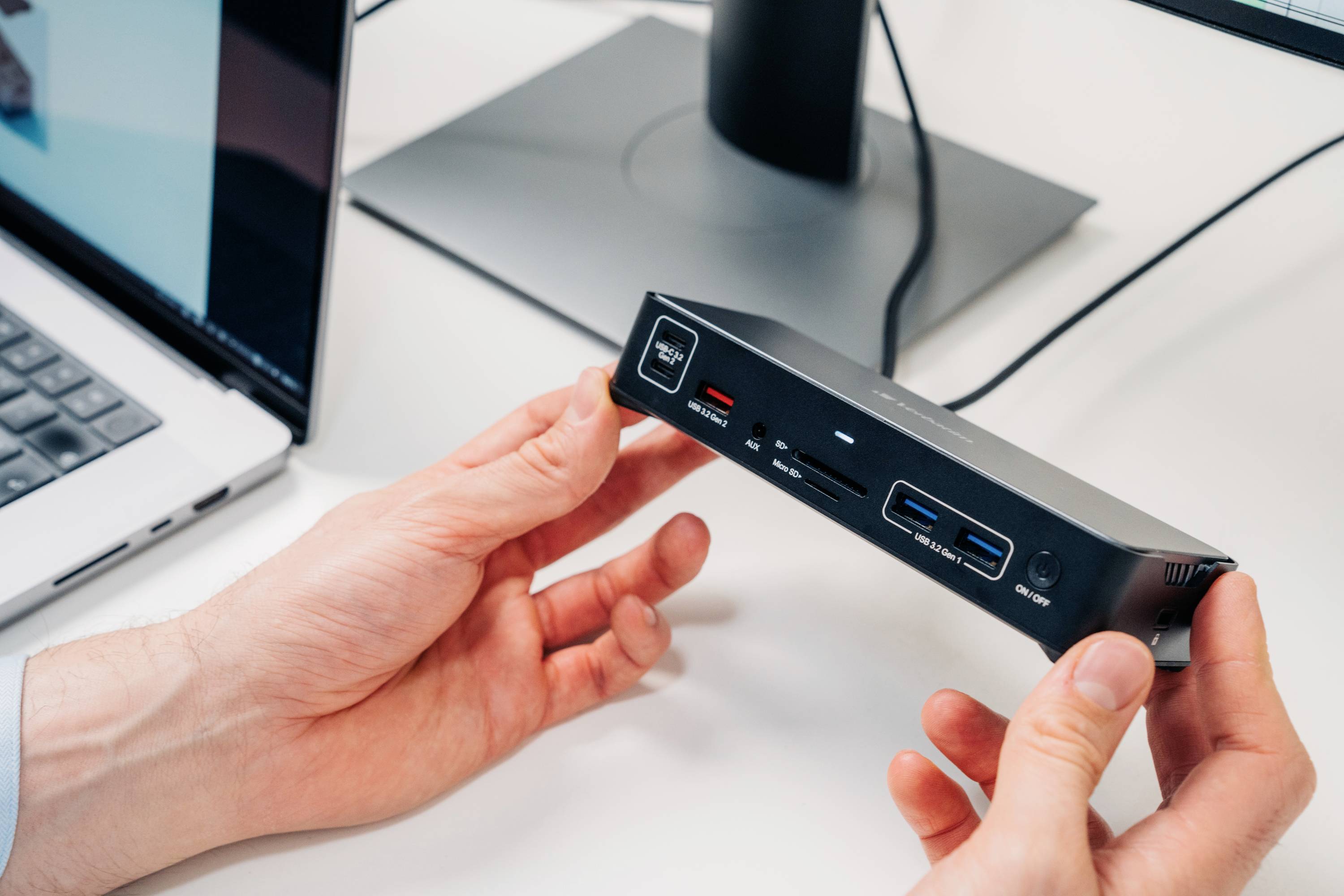 Een persoon houdt een elektronisch apparaat met meerdere aansluitingen, waaronder USB- en HDMI-poorten, in de hand. Op de achtergrond een laptop.