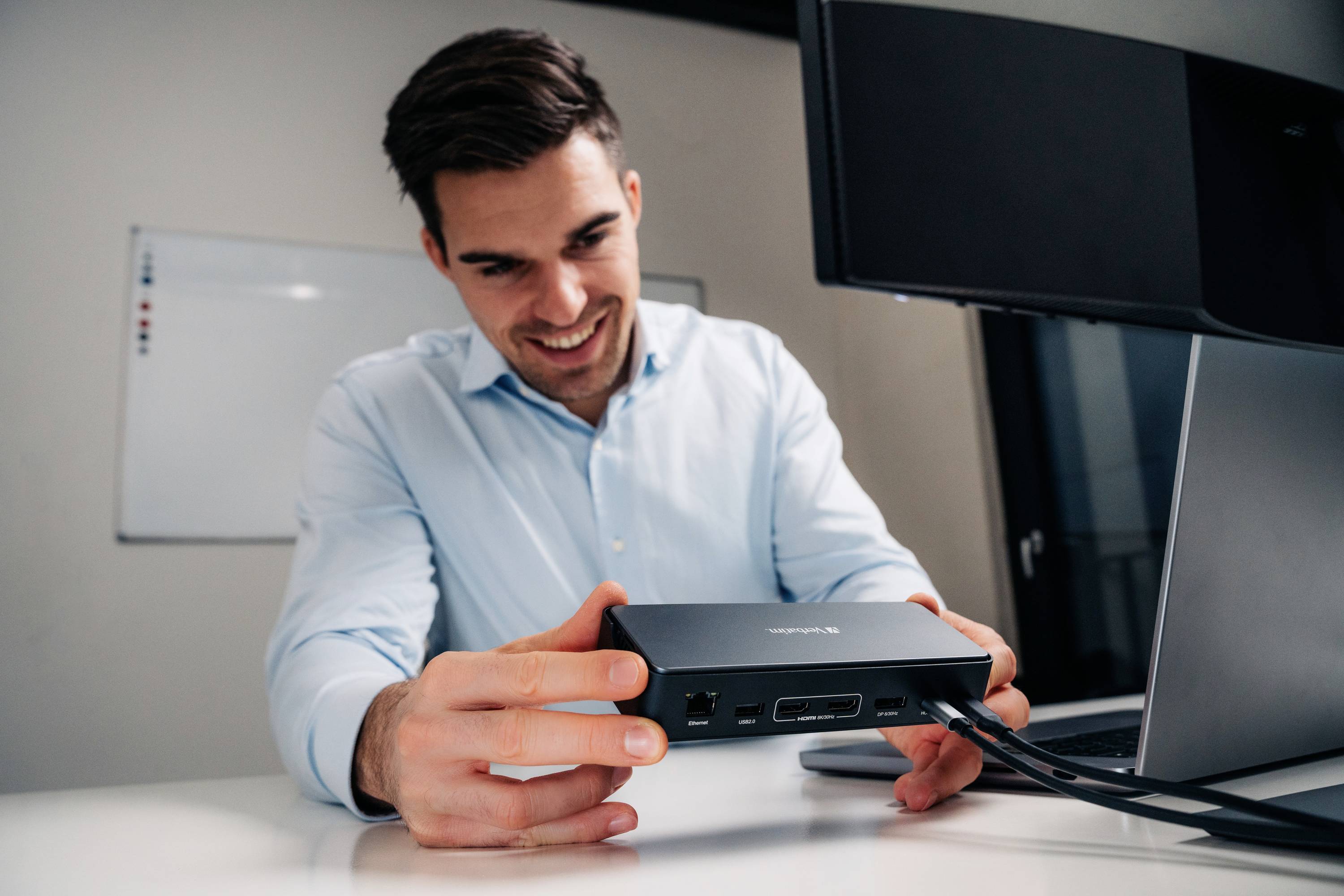 Een man in een lichtblauw overhemd zit aan een bureau en glimlacht terwijl hij een elektronisch apparaat met meerdere aansluitingen vasthoudt. Op de achtergrond zijn een monitor en een whiteboard zichtbaar.