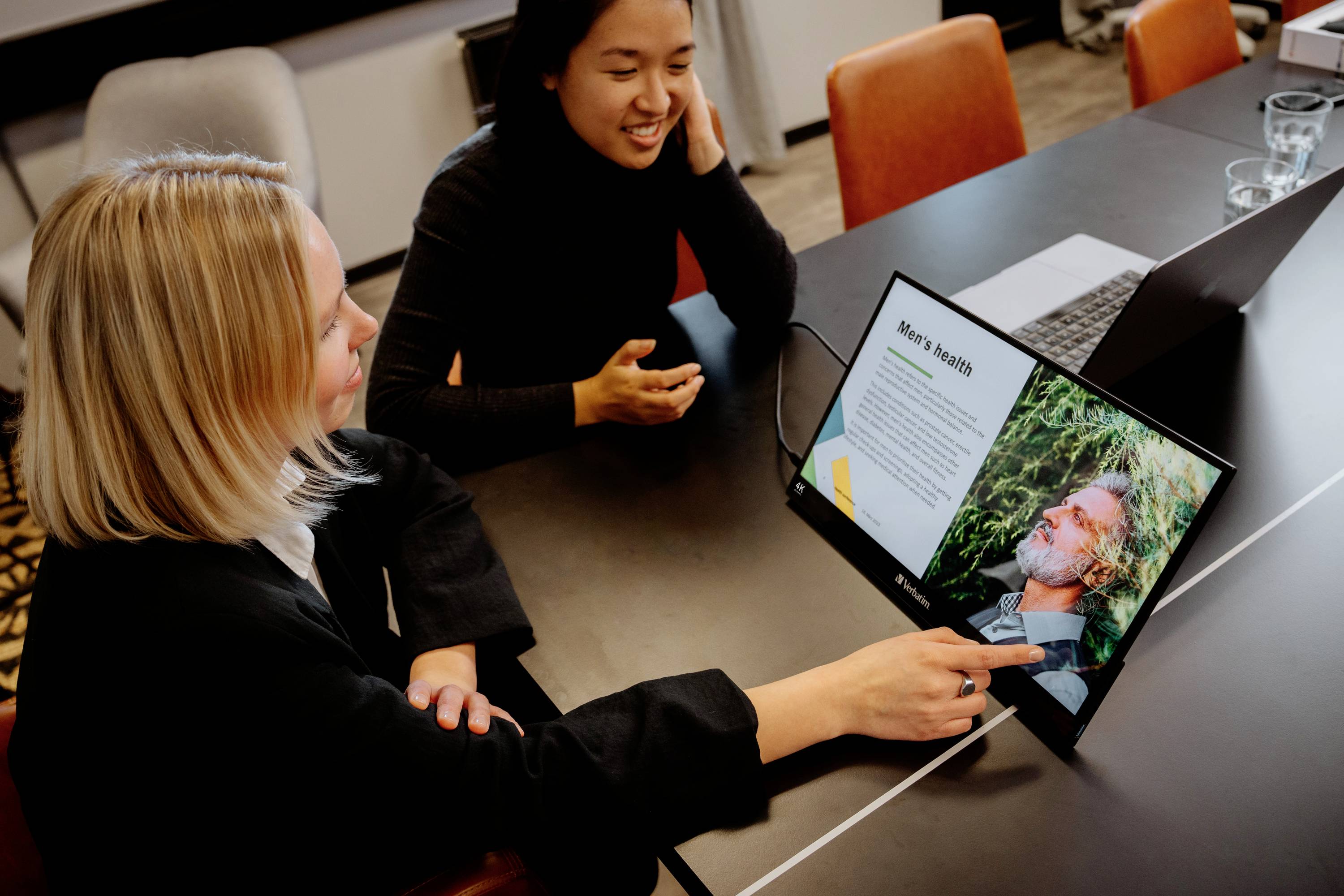 Twee personen in een vergaderruimte bekijken samen het scherm van een laptop met een presentatie over 'Herentrends'.