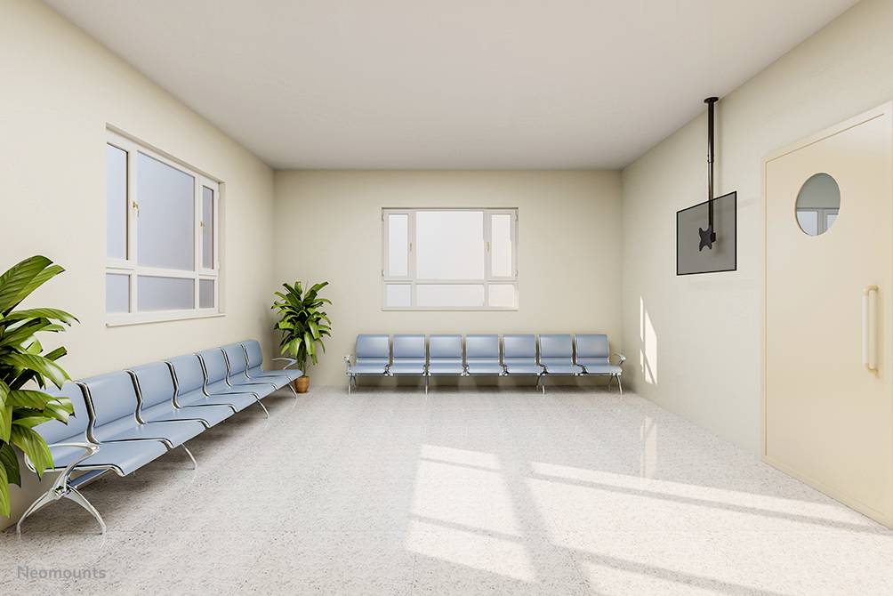 Een lege wachtkamer met blauwe stoelen, twee planten en een televisie aan de muur. Twee grote ramen laten zonlicht naar binnen.