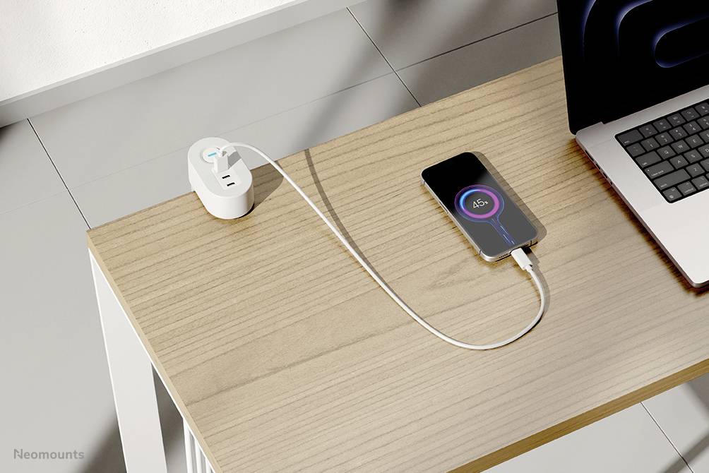 Een smartphone wordt opgeladen op een houten tafel naast een laptop; hij is verbonden met een wit kabel dat in een stopcontact steekt.