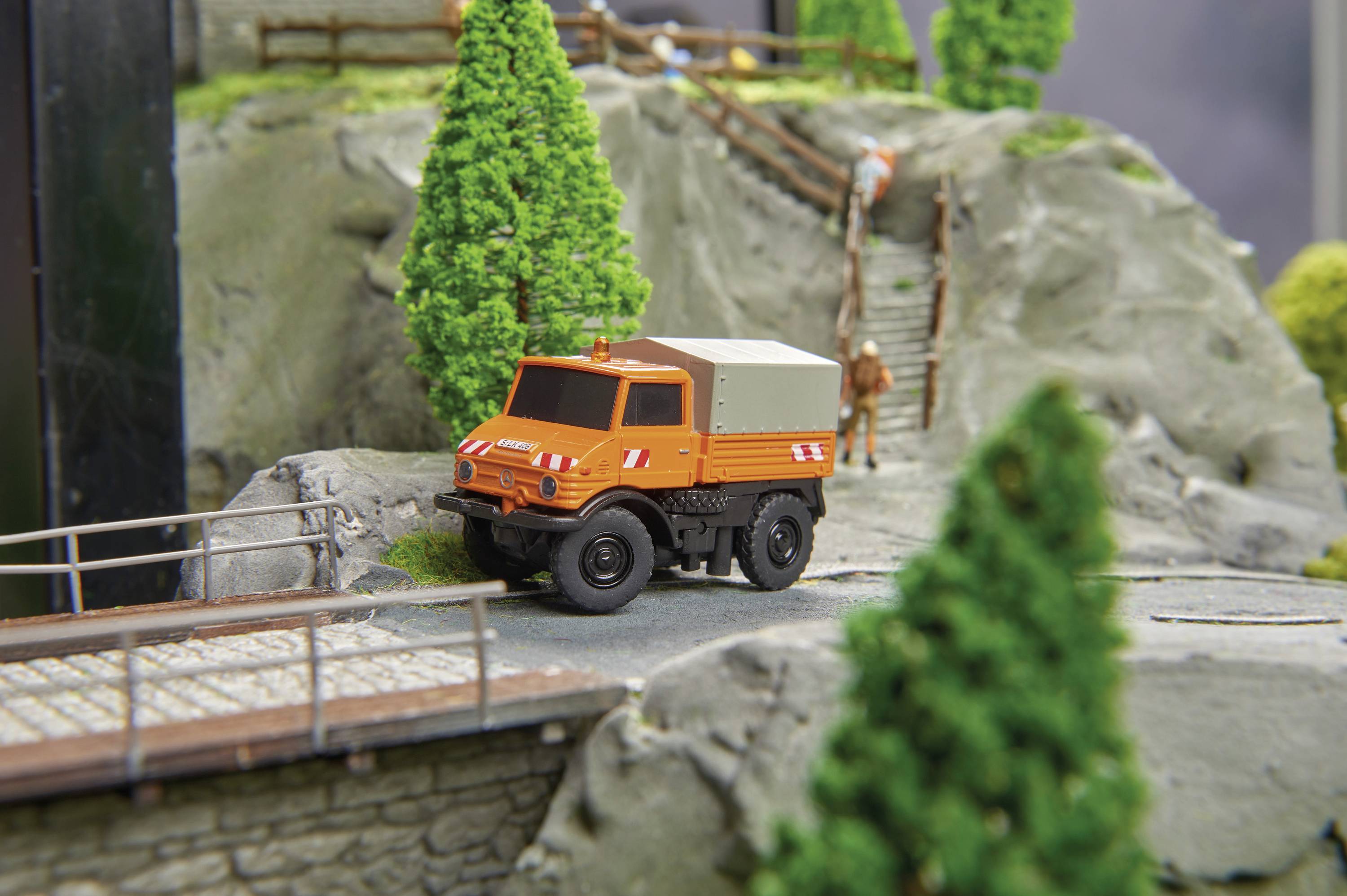 Oranje modelvoertuig op een miniatuurlandschap met bomen en rotsen, op de achtergrond staan figuren op een trap.