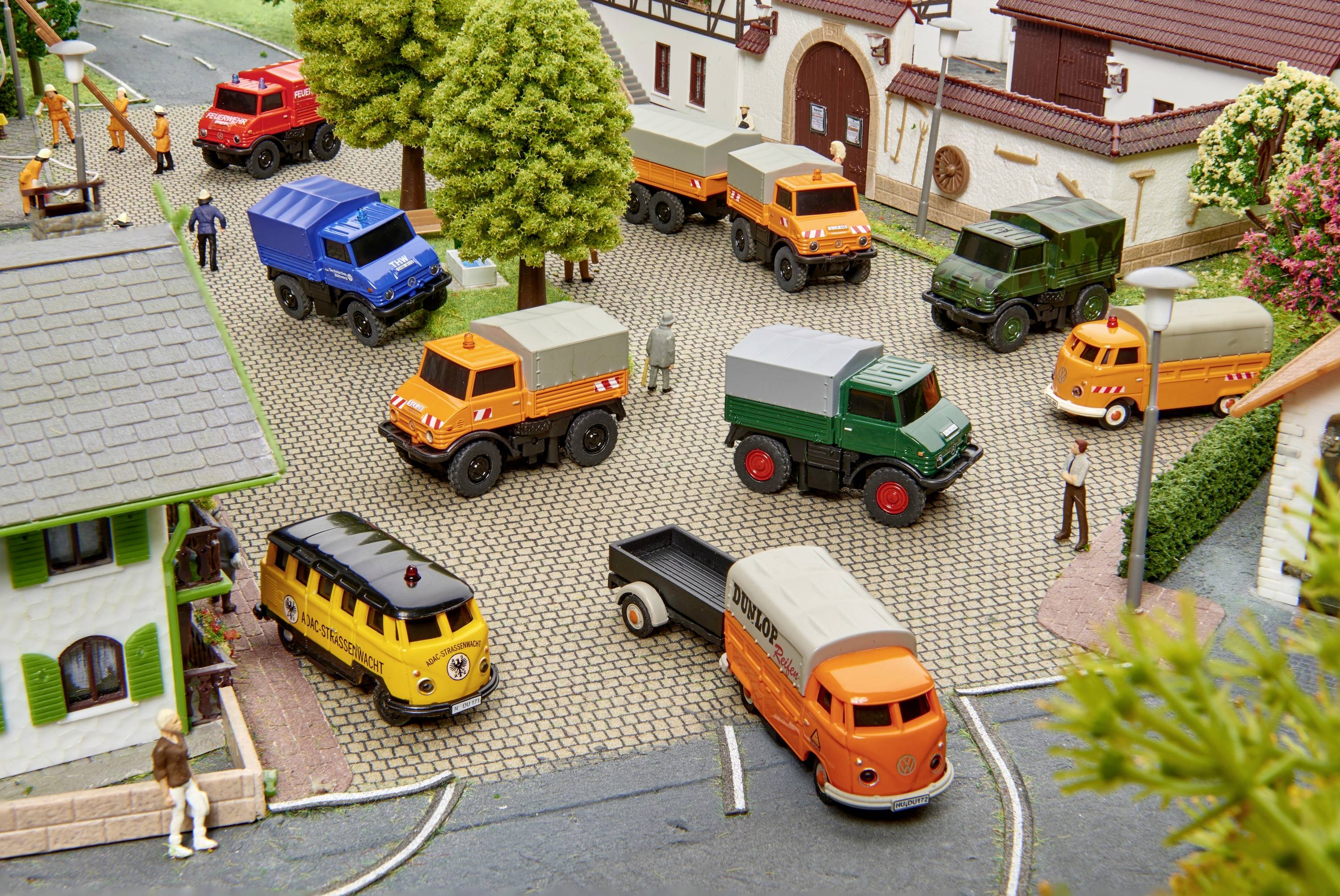 Een miniatuurscène toont kleurrijke modelauto's en -vrachtwagens op een geplaveid plein, omringd door gebouwen en bomen. Menselijke figuren staan verspreid.
