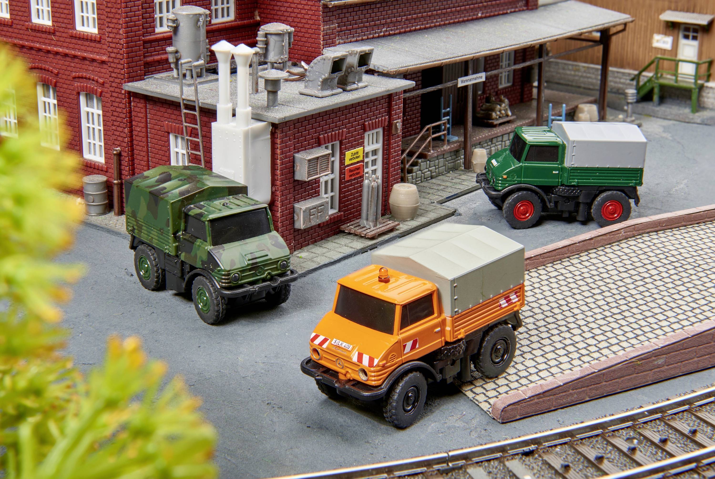 Drie model-vrachtwagens parkeren voor een gebouw in een miniatuuropstelling. Eén is oranje, een andere is groen gecamoufleerd, en de derde is groen.