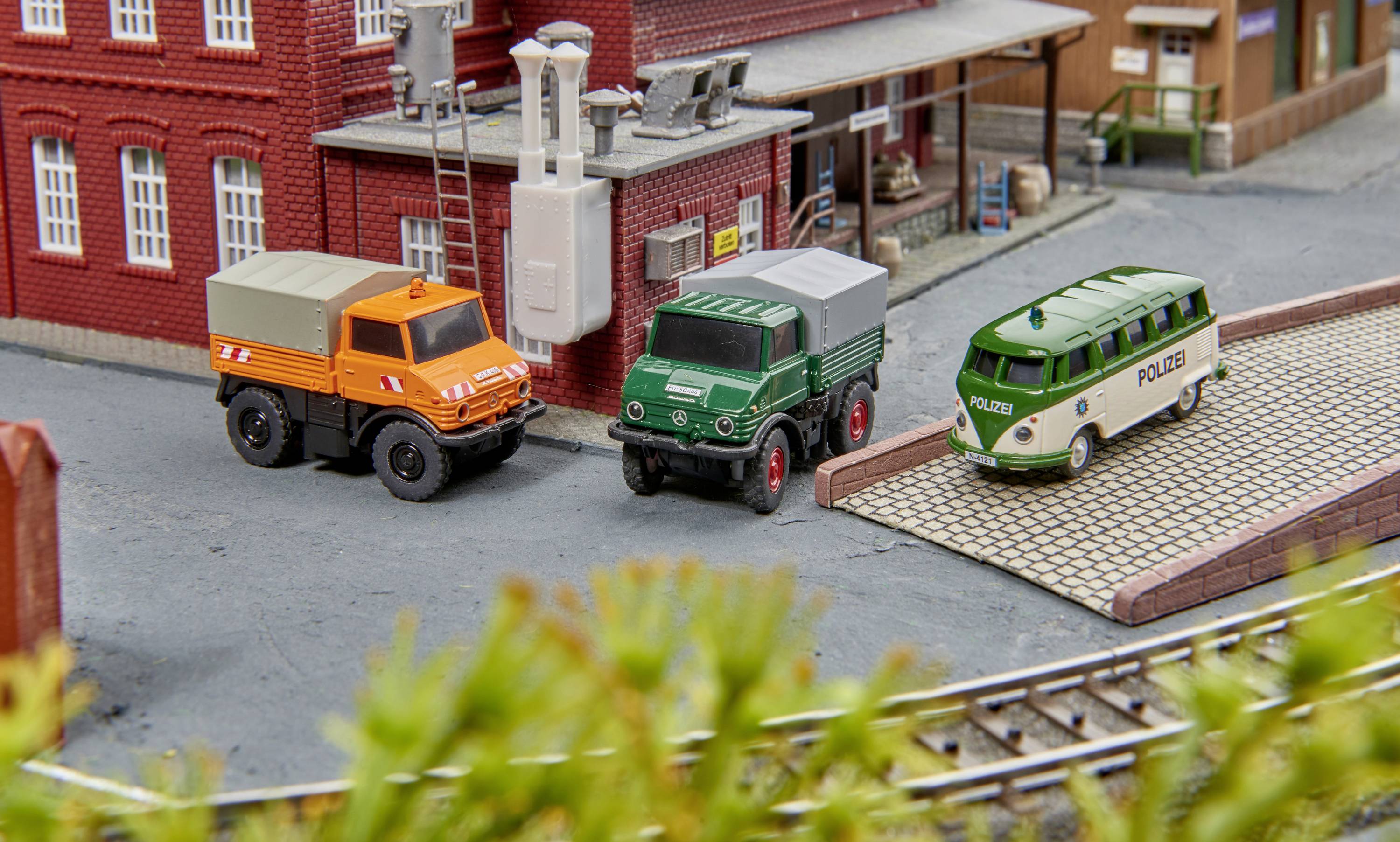 Drie modelvoertuigen, waaronder een oranje en een groene vrachtwagen evenals een groene politiebus, voor een modelgebouw.