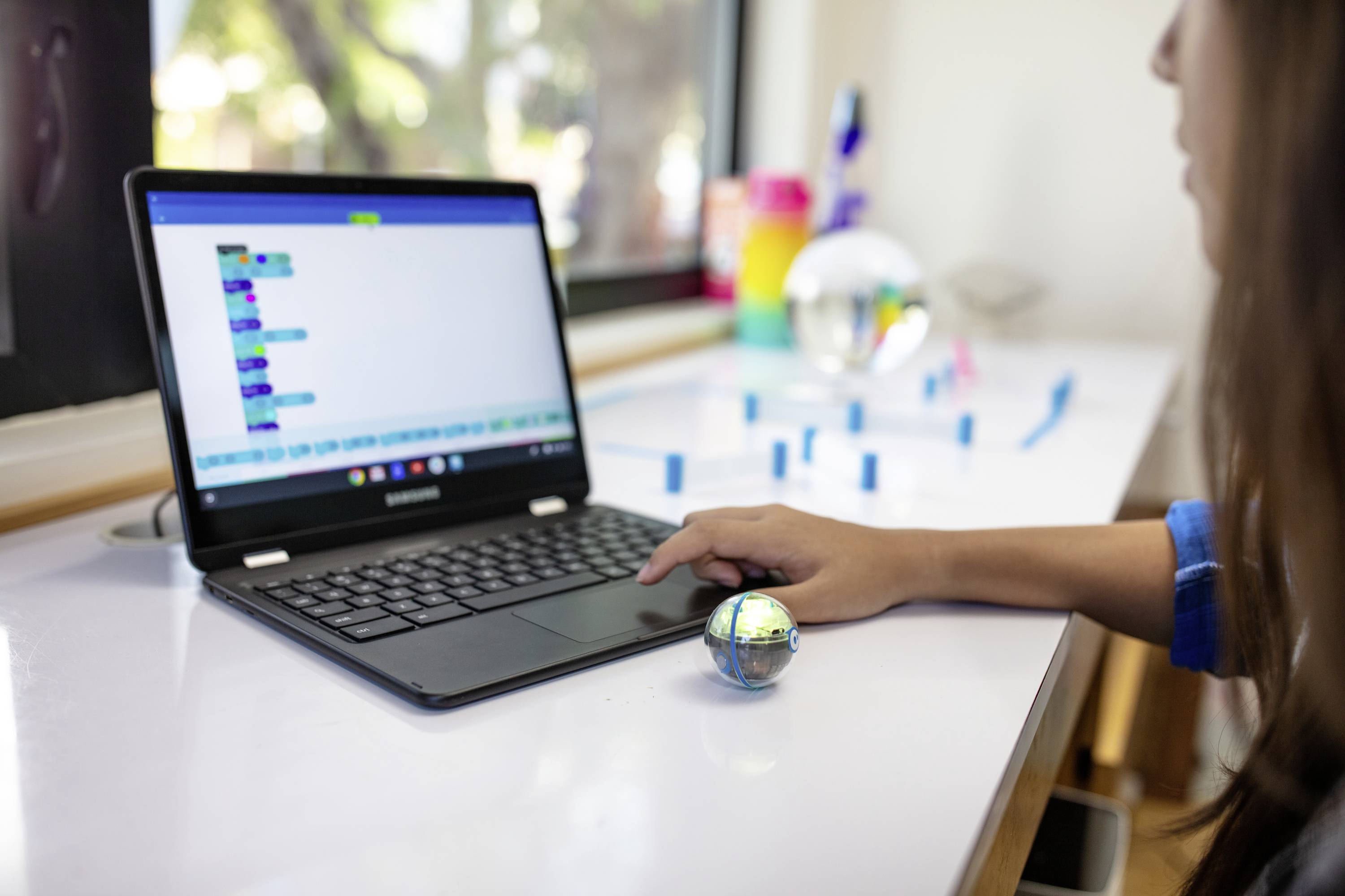 Een kind programmeert een kleine robotbal met een laptop. Op het scherm zijn kleurrijke programmeerblokken te zien.