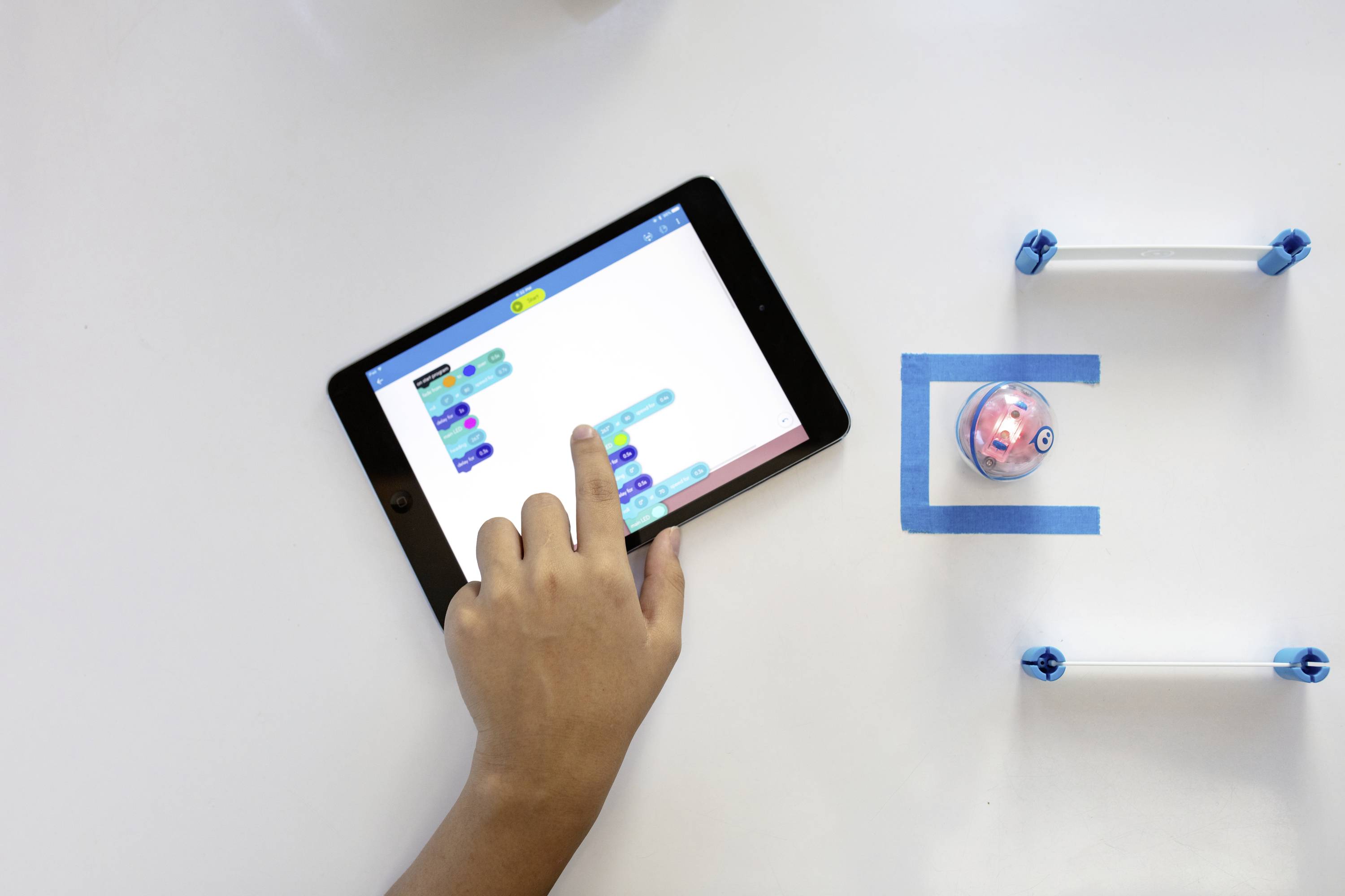 Hand bedient tablet met programmeeromgeving, naast kleine robot op tafel. Robotica en programmering in onderwijsomgeving.