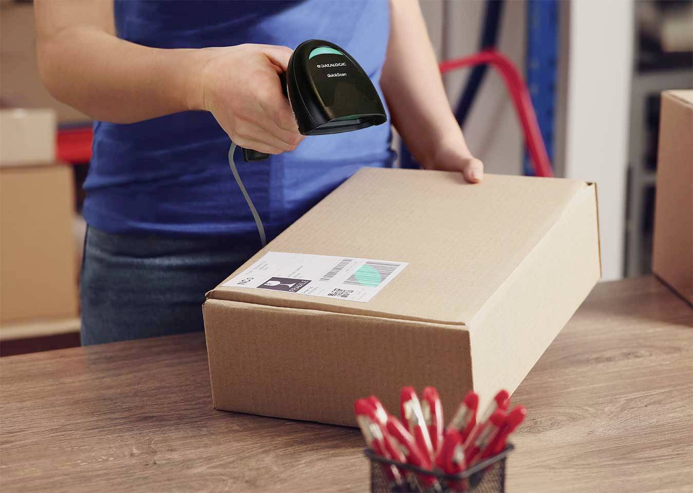 Een persoon scant een verzendlabel op een kartonnen doos met een handscanner op een tafel. Op de voorgrond staat een bak met rode pennen.