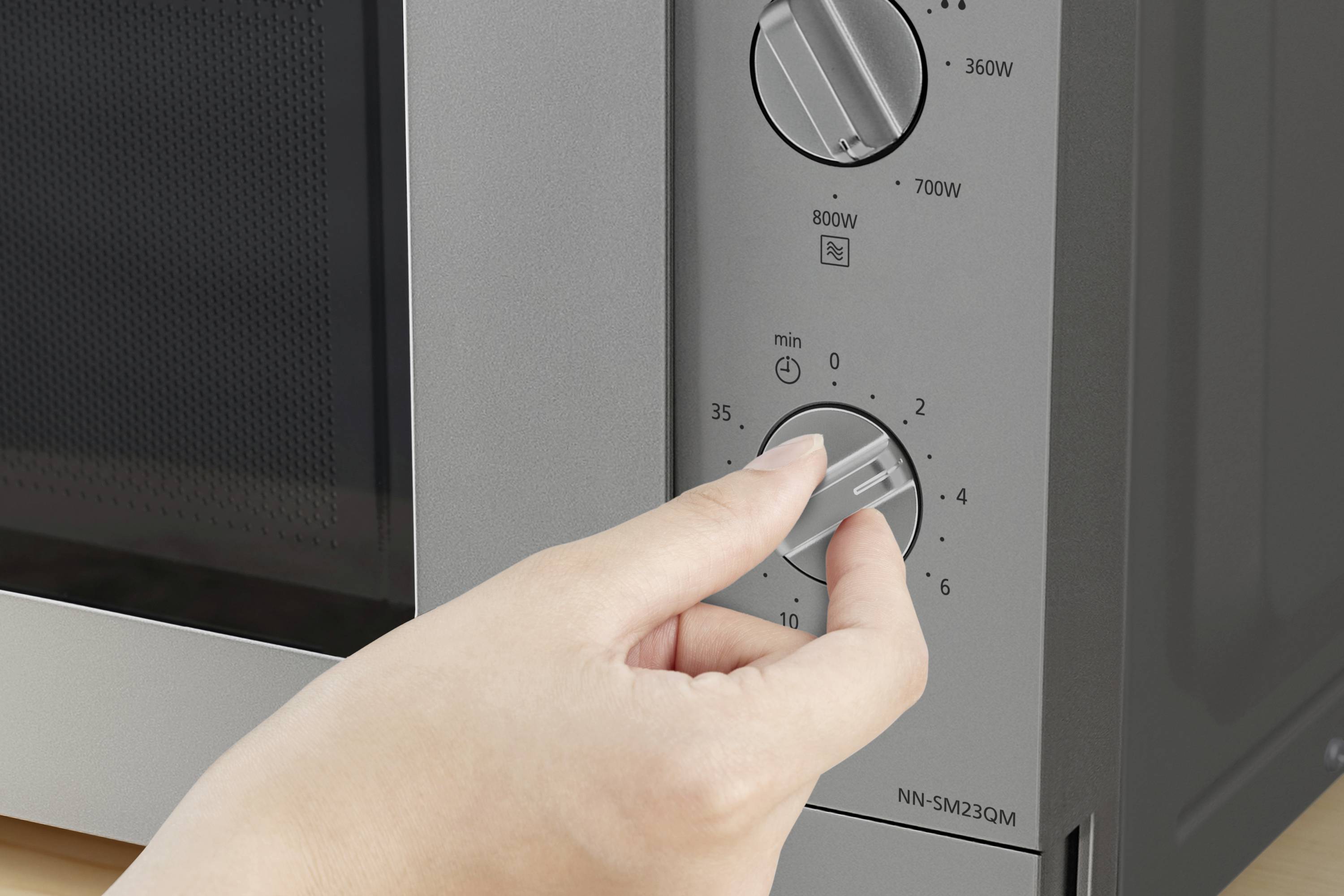 Een hand draait aan de tijdschakelaar van een magnetron, die wordt ingesteld in minuten. Twee andere draaiknoppen zijn bedoeld voor de vermogensinstellingen.