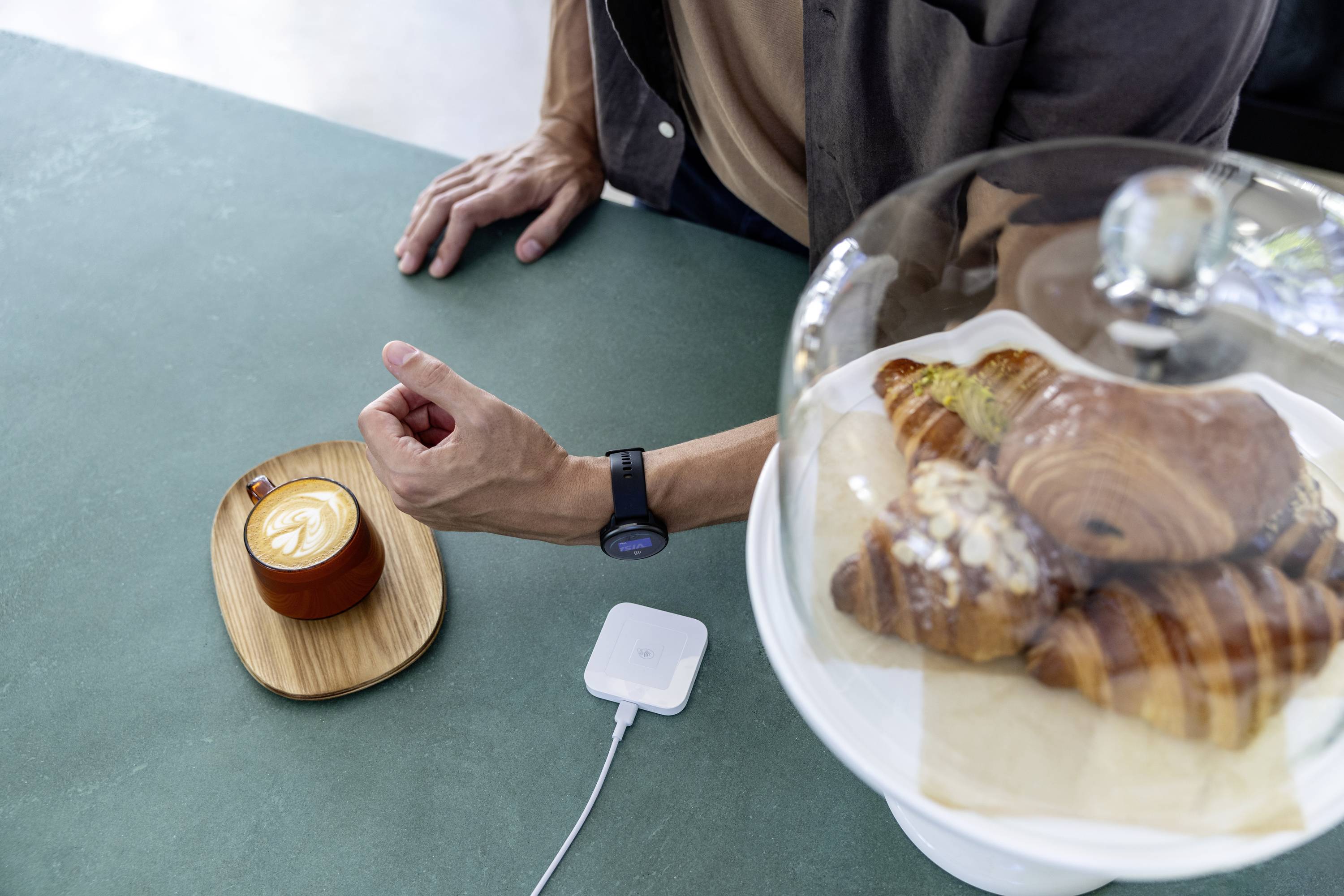 Een persoon wijst naar een cappuccino op een houten bord naast een glazen stolp met croissants op een groene tafel.