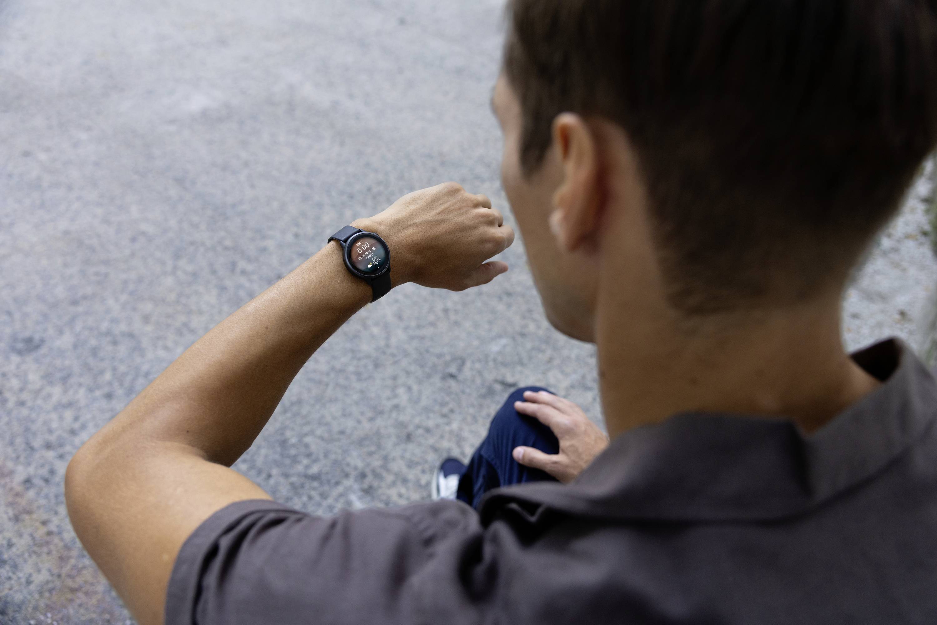 Een persoon kijkt naar een smartwatch aan de pols. De omgeving is een grijze, asfalterende oppervlakte.