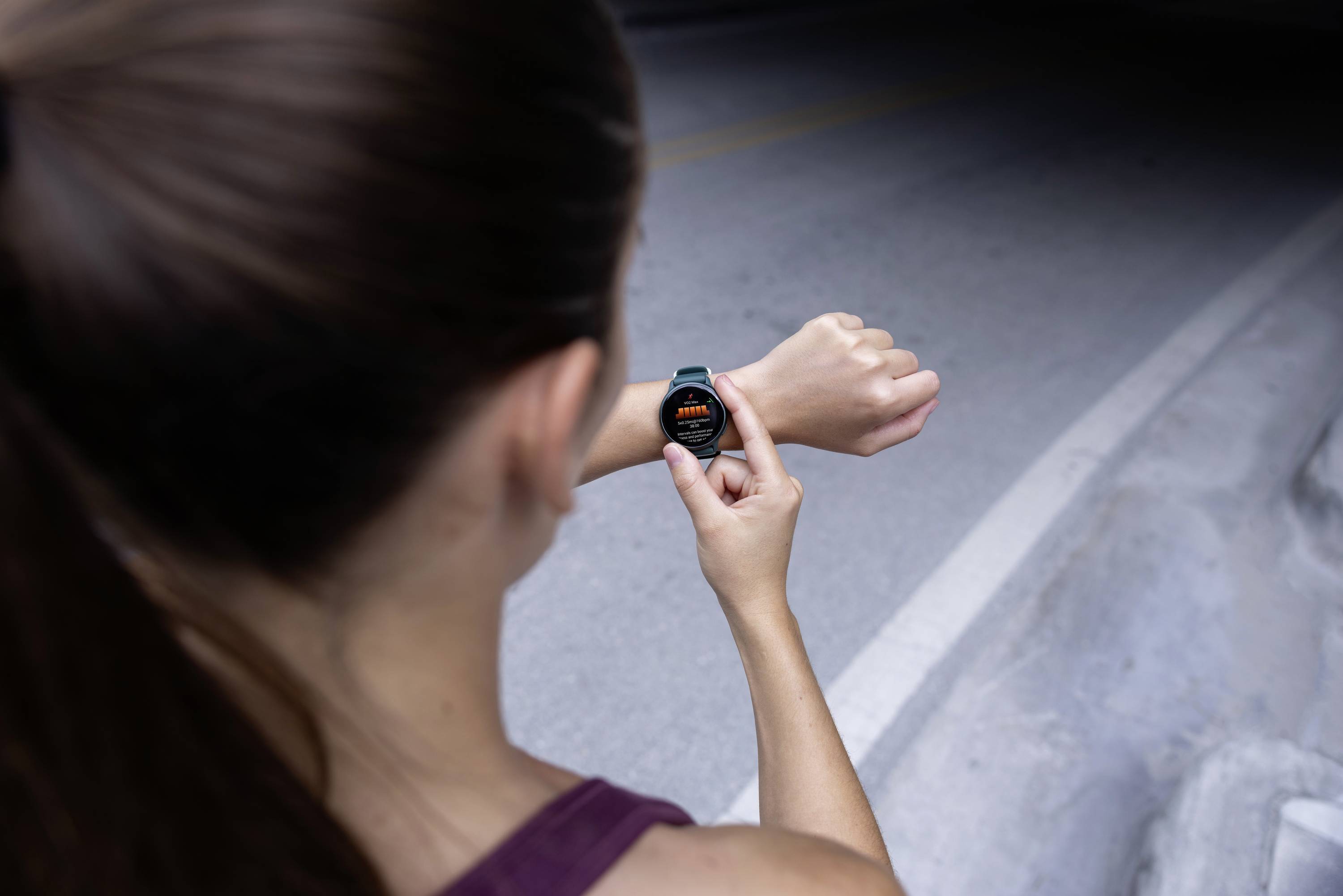 Een vrouw controleert buiten een smartwatch die stappen weergeeft. Ze draagt sportkleding en staat op een geasfalteerd pad.