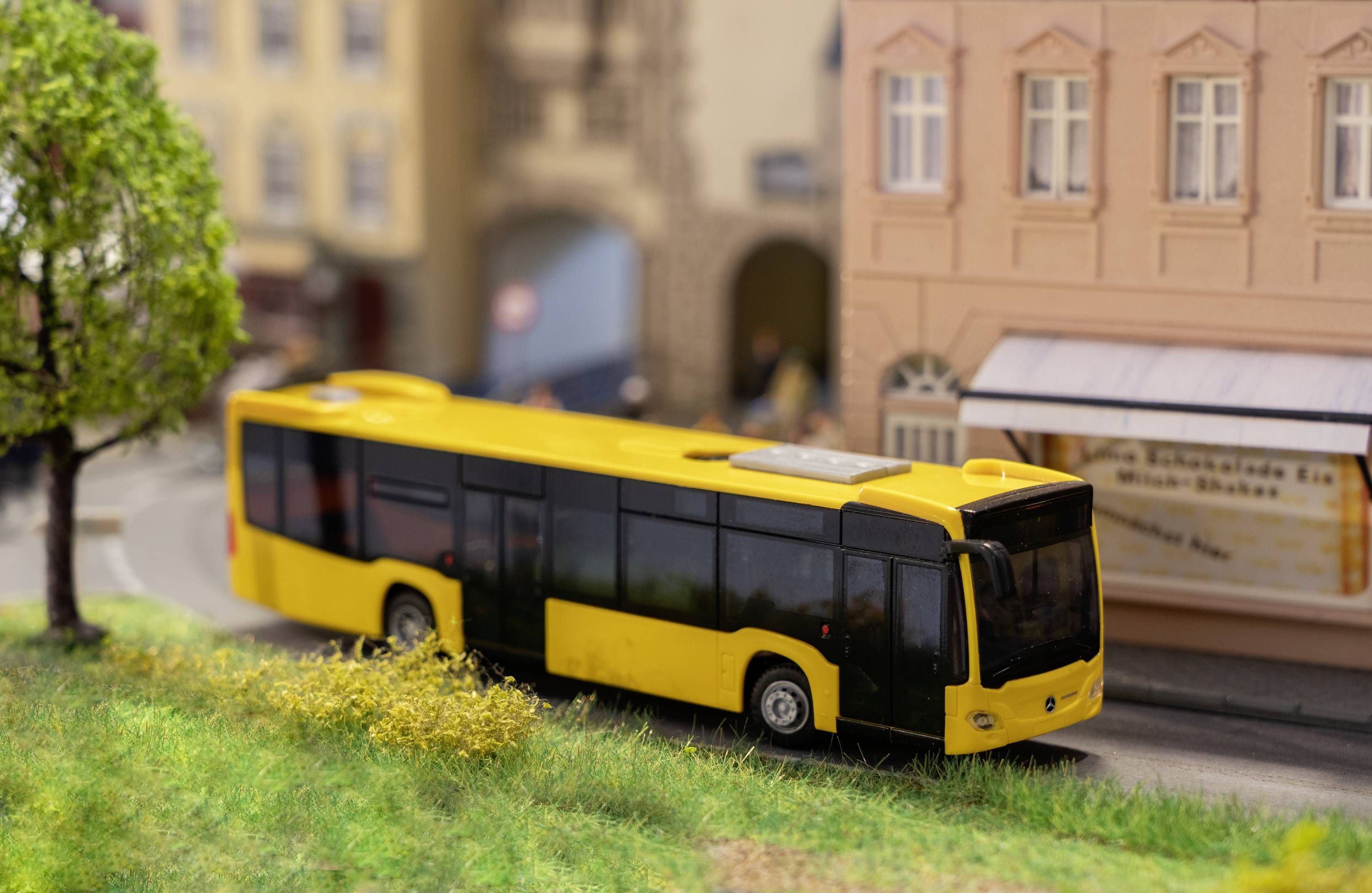 Een gele modelbus rijdt op een miniatuurweg naast gebouwen, een boom en een groenstrook.