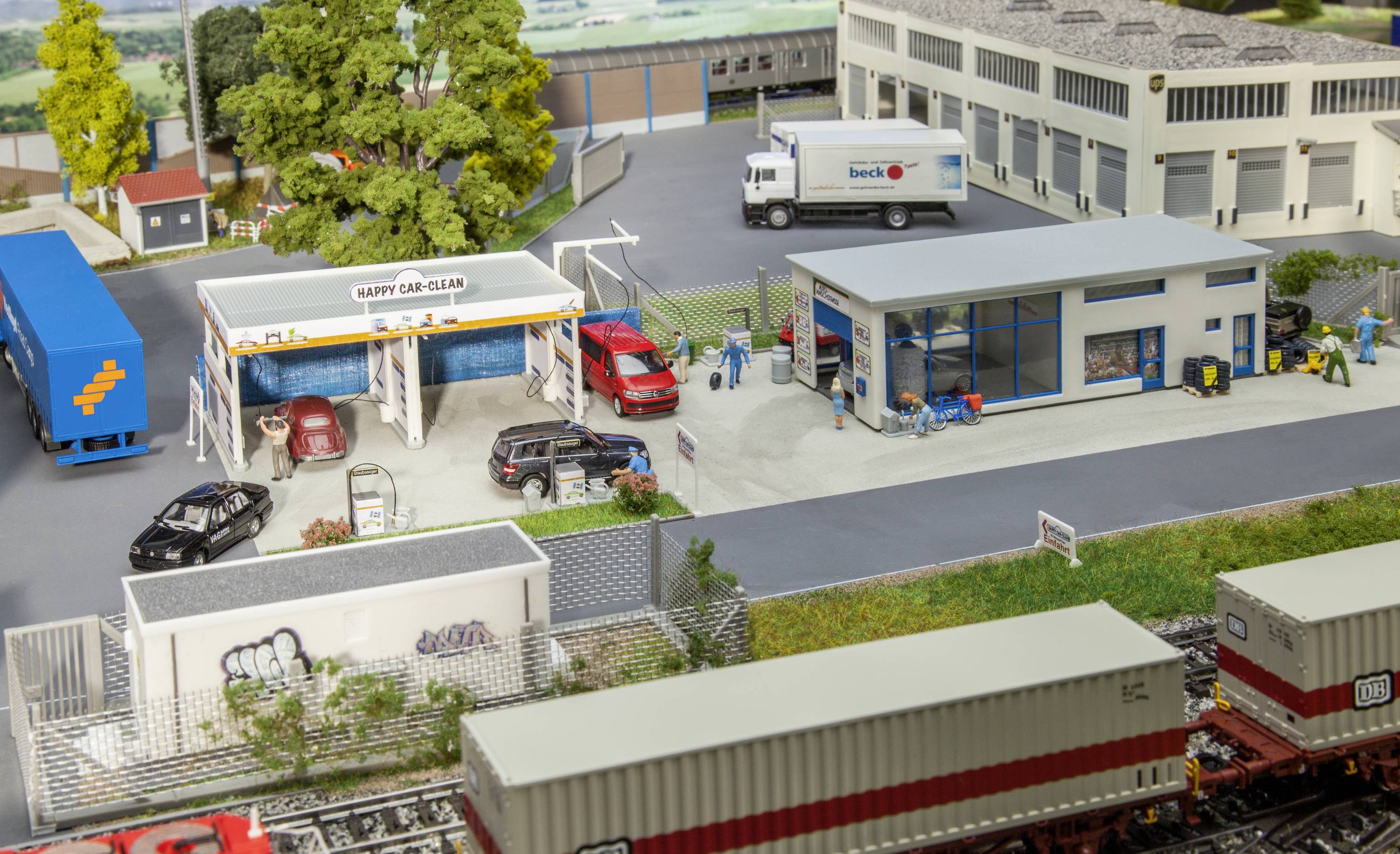 Miniatuur van een autowasstraat en werkplaats met auto's en figuren. Naast de wasstraat bevinden zich gebouwen, vrachtwagens en containers in een modelandschap.