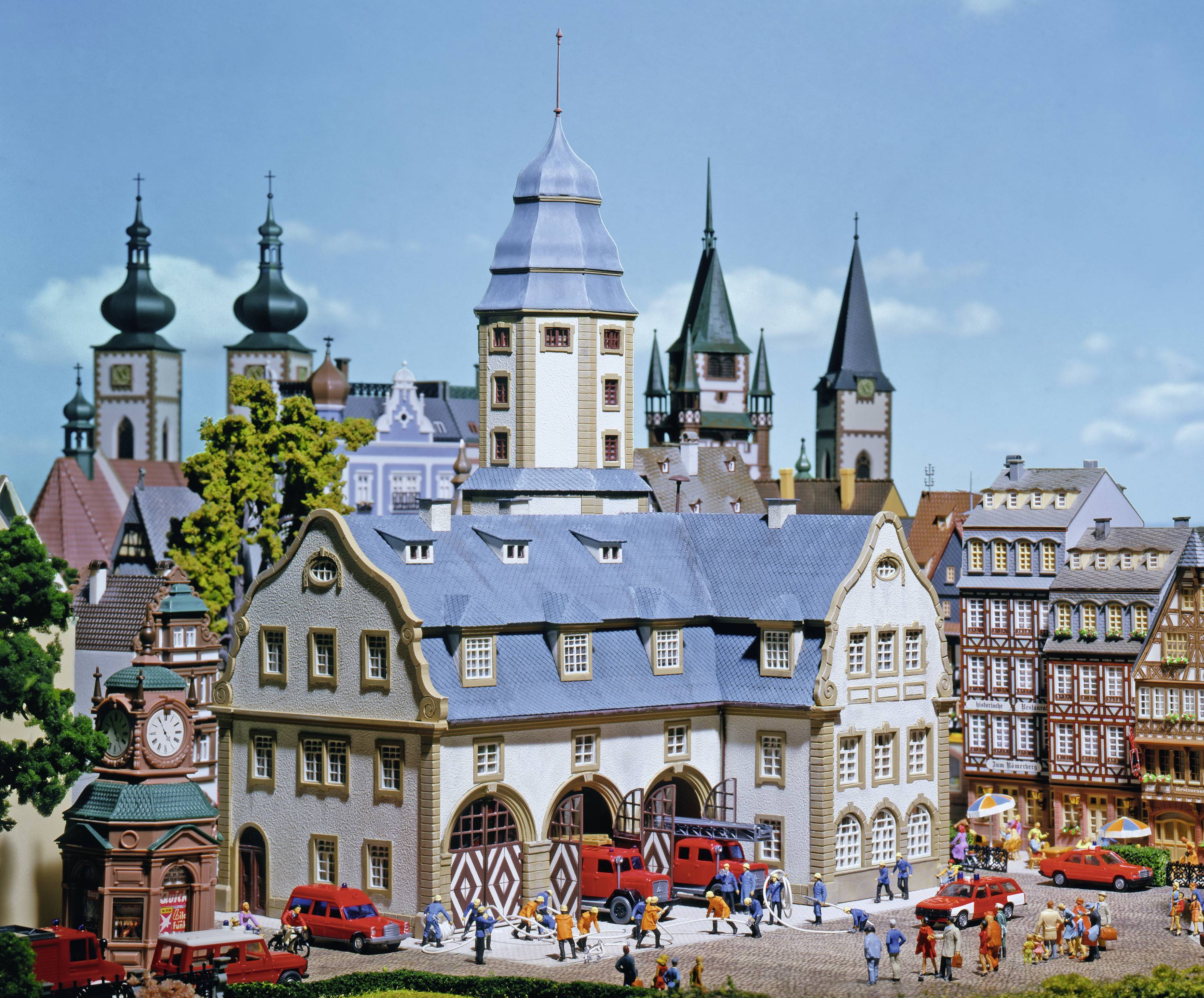 Modelstad met historische gebouwen, kerktorens en kleine figuren die over de markt wandelen. Auto's parkeren in de buurt.