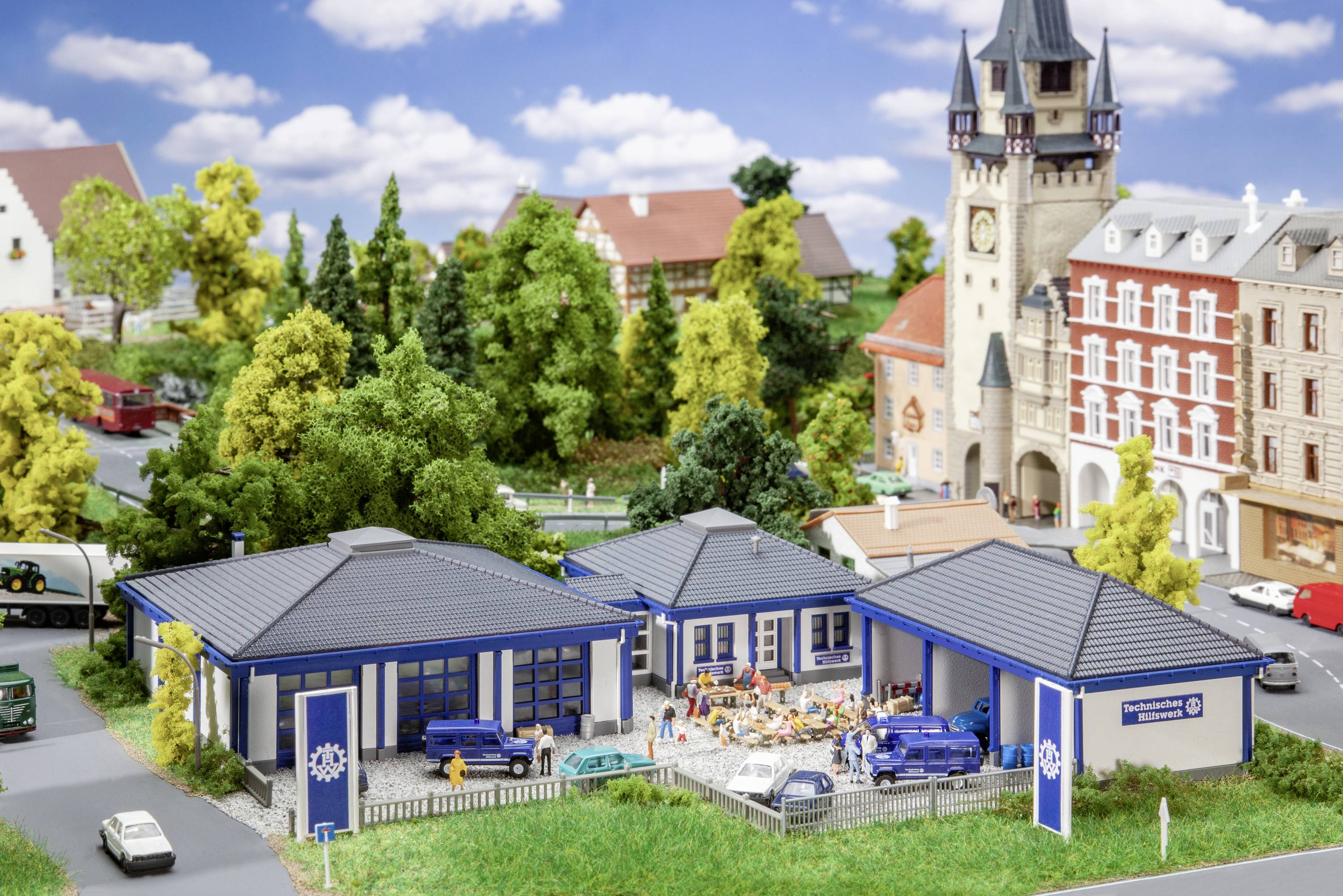 Modelspoorbaan landschap met gebouwen en voertuigen, waaronder een brandweerwagen. Op de achtergrond een toren en huizen, omgeven door bomen.